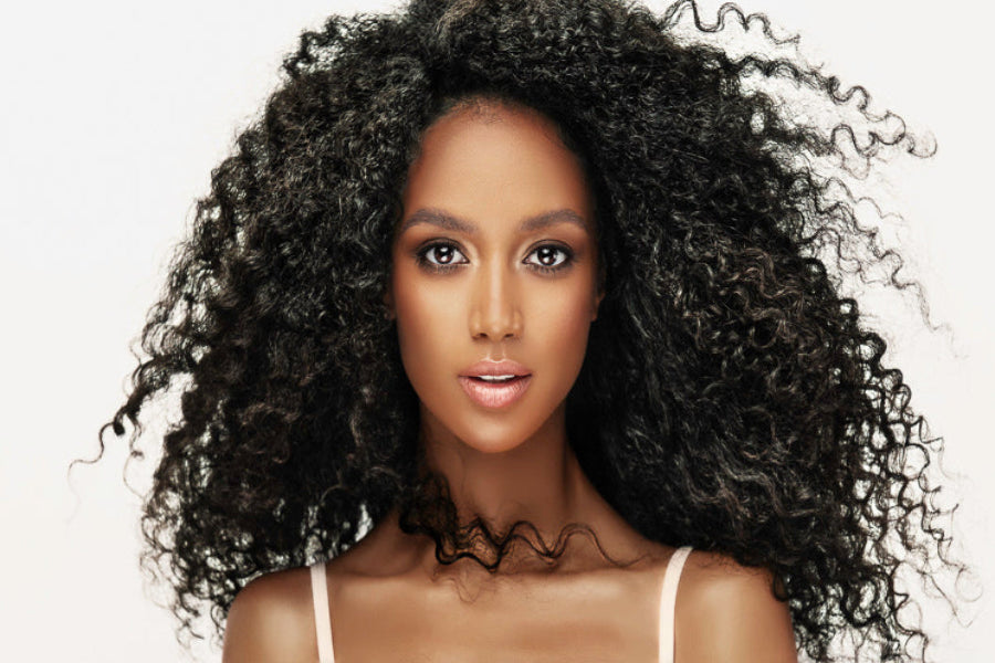 A close-up portrait of a woman with voluminous, tightly curled black hair, wearing a light-colored spaghetti strap top against a plain white background.